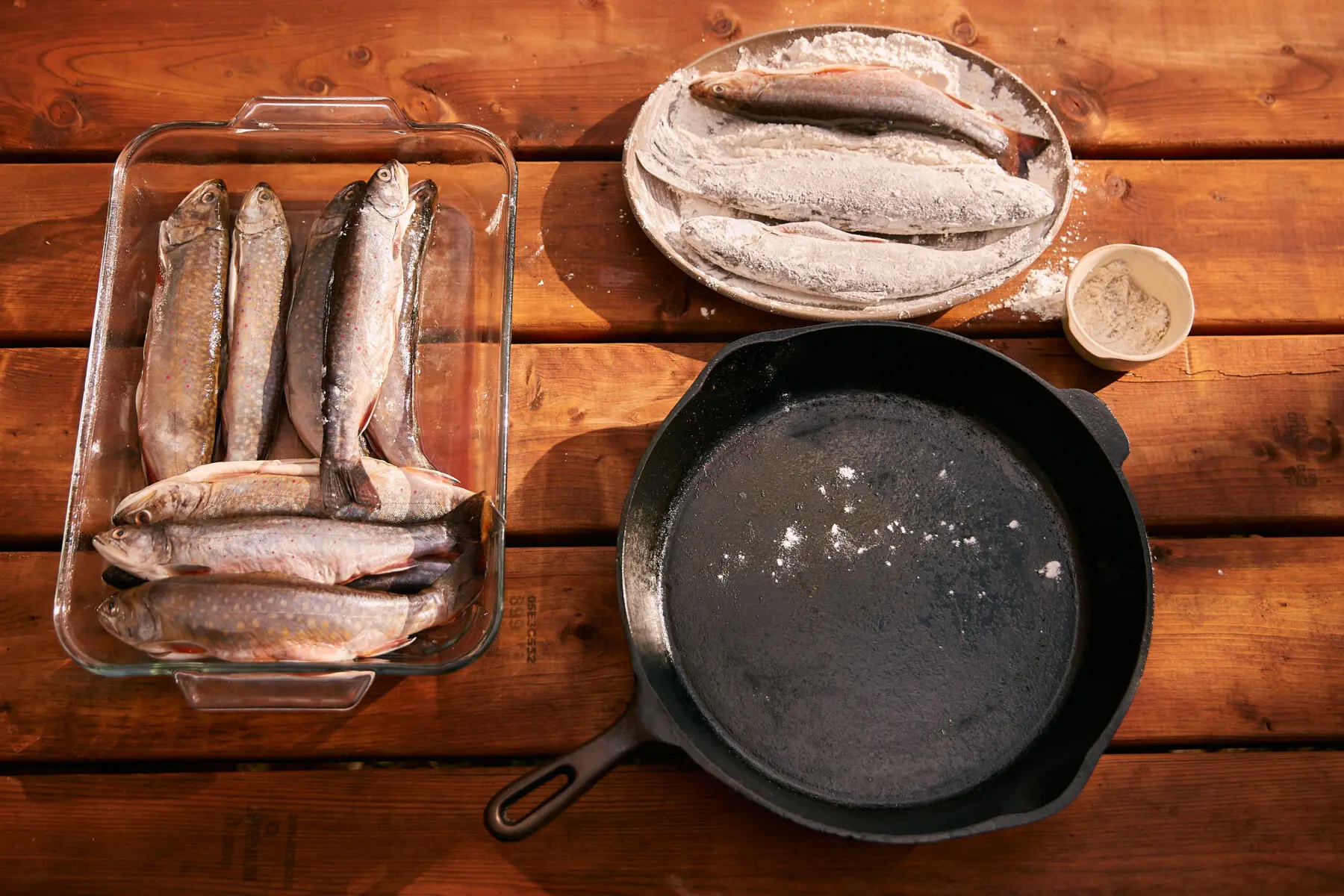 Fresh trout salted in a dish, floured fillets and a cast iron skillet ready for fire grilled fish