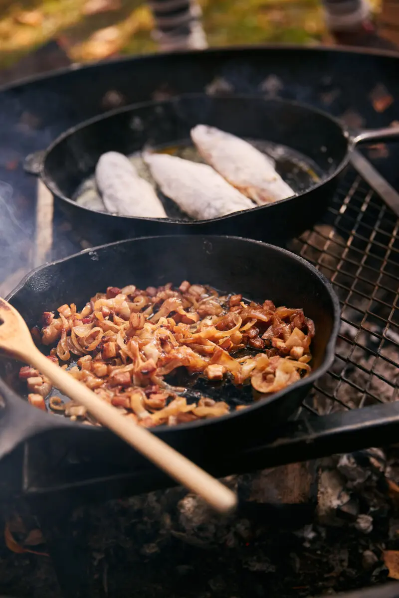 Bacon and onions frying in a cast iron skillet beside floured trout fillets over the campfire