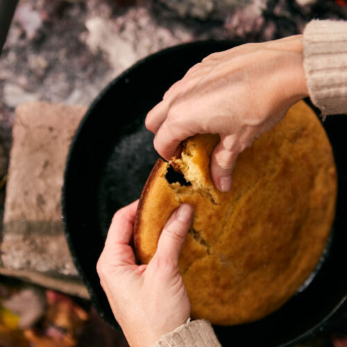 Hands pulling apart golden cast iron cornbread fresh from a campfire Dutch oven