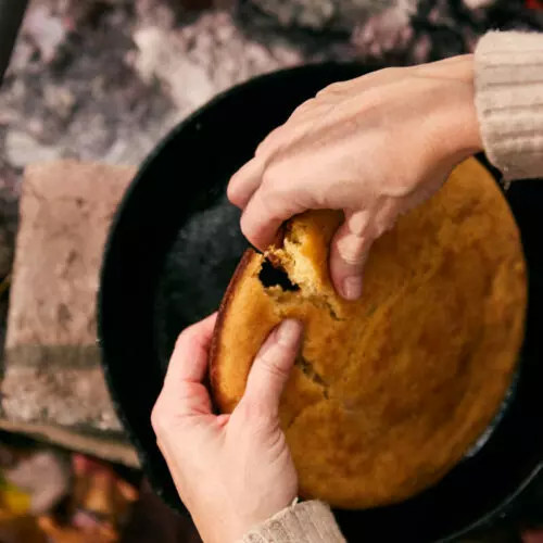 Hands pulling apart golden cast iron cornbread fresh from a campfire Dutch oven