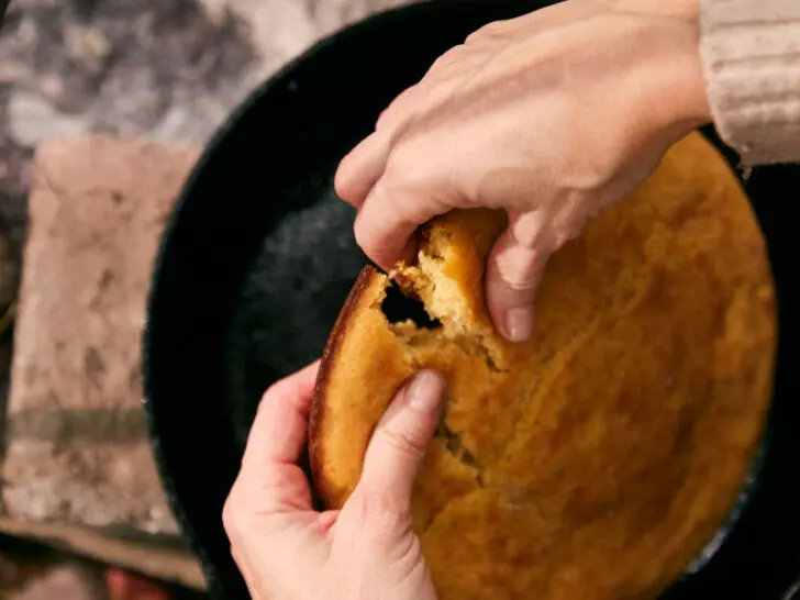 Hands pulling apart golden cast iron cornbread fresh from a campfire Dutch oven