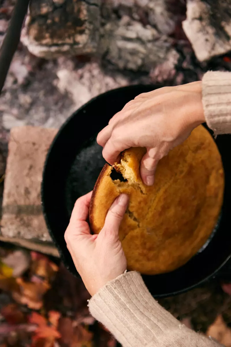 Hands pulling apart golden cast iron cornbread fresh from a campfire Dutch oven