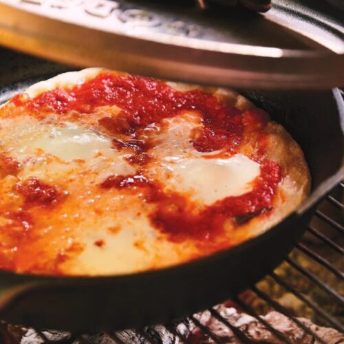 Cast iron skillet pizza with bubbling cheese cooking over hot campfire coals