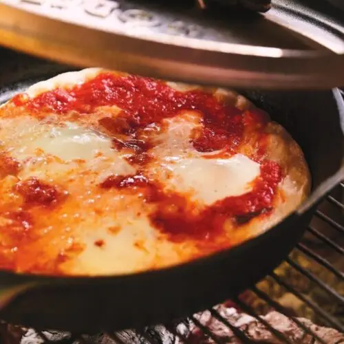 Cast iron skillet pizza with bubbling cheese cooking over hot campfire coals