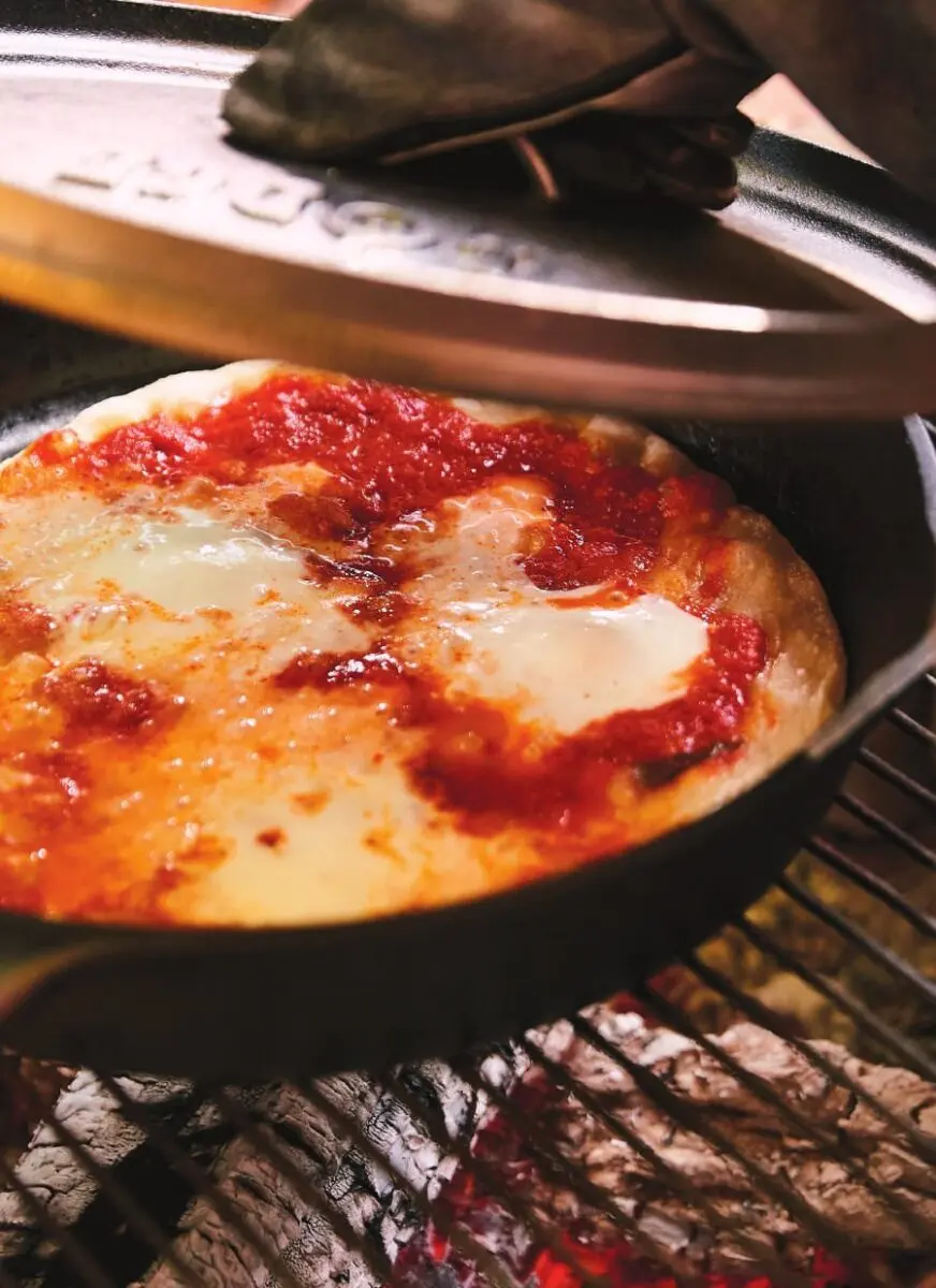 Cast iron skillet pizza with bubbling cheese cooking over hot campfire coals
