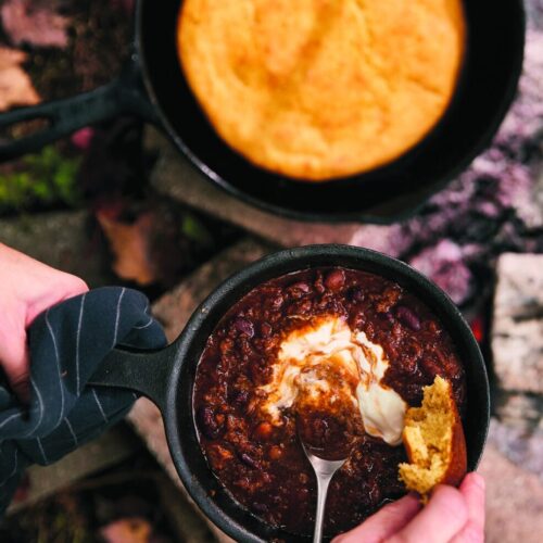 Campfire chili in a cast iron skillet with cornbread and sour cream by the fire
