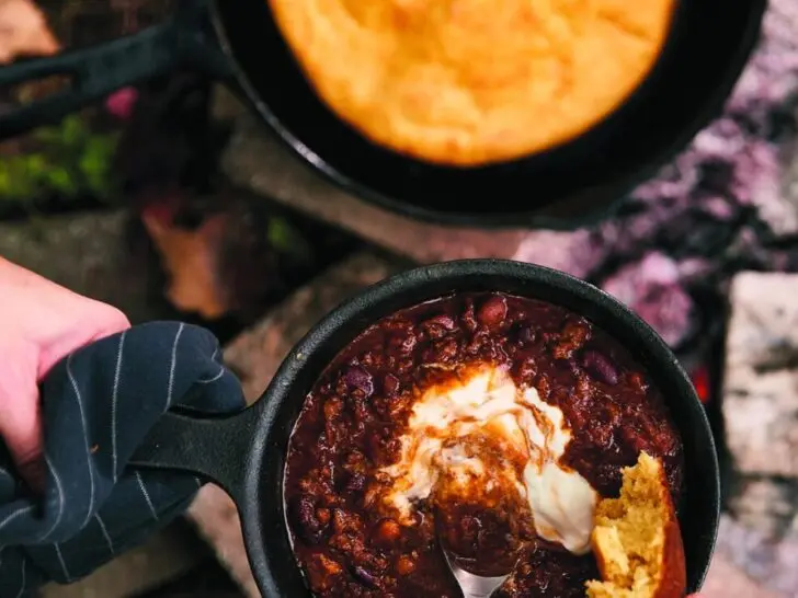 Campfire chili in a cast iron skillet with cornbread and sour cream by the fire