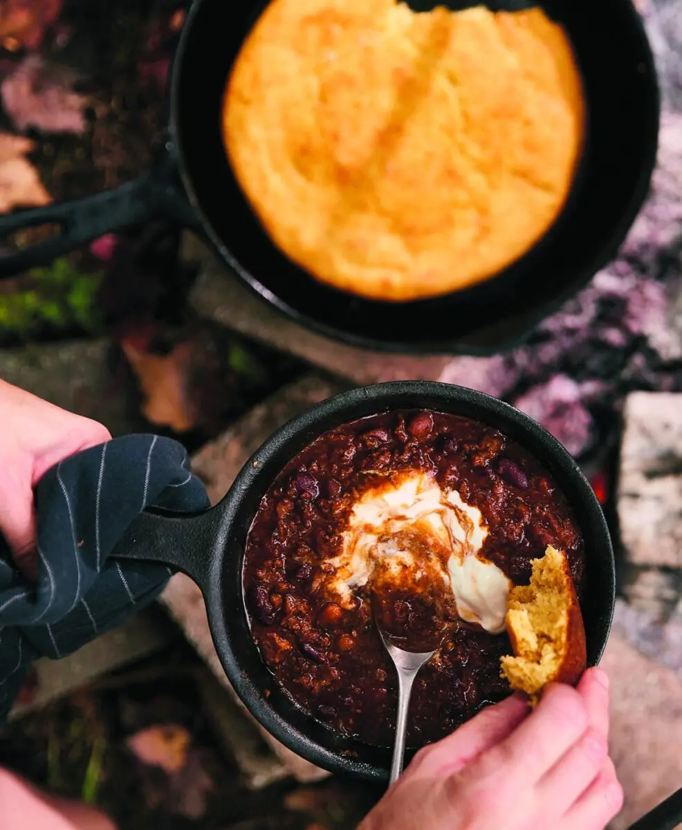 Campfire chili in a cast iron skillet with cornbread and sour cream by the fire