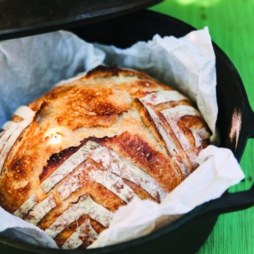 Golden dutch oven bread with a scored crust lifting out of a cast iron pot on parchment
