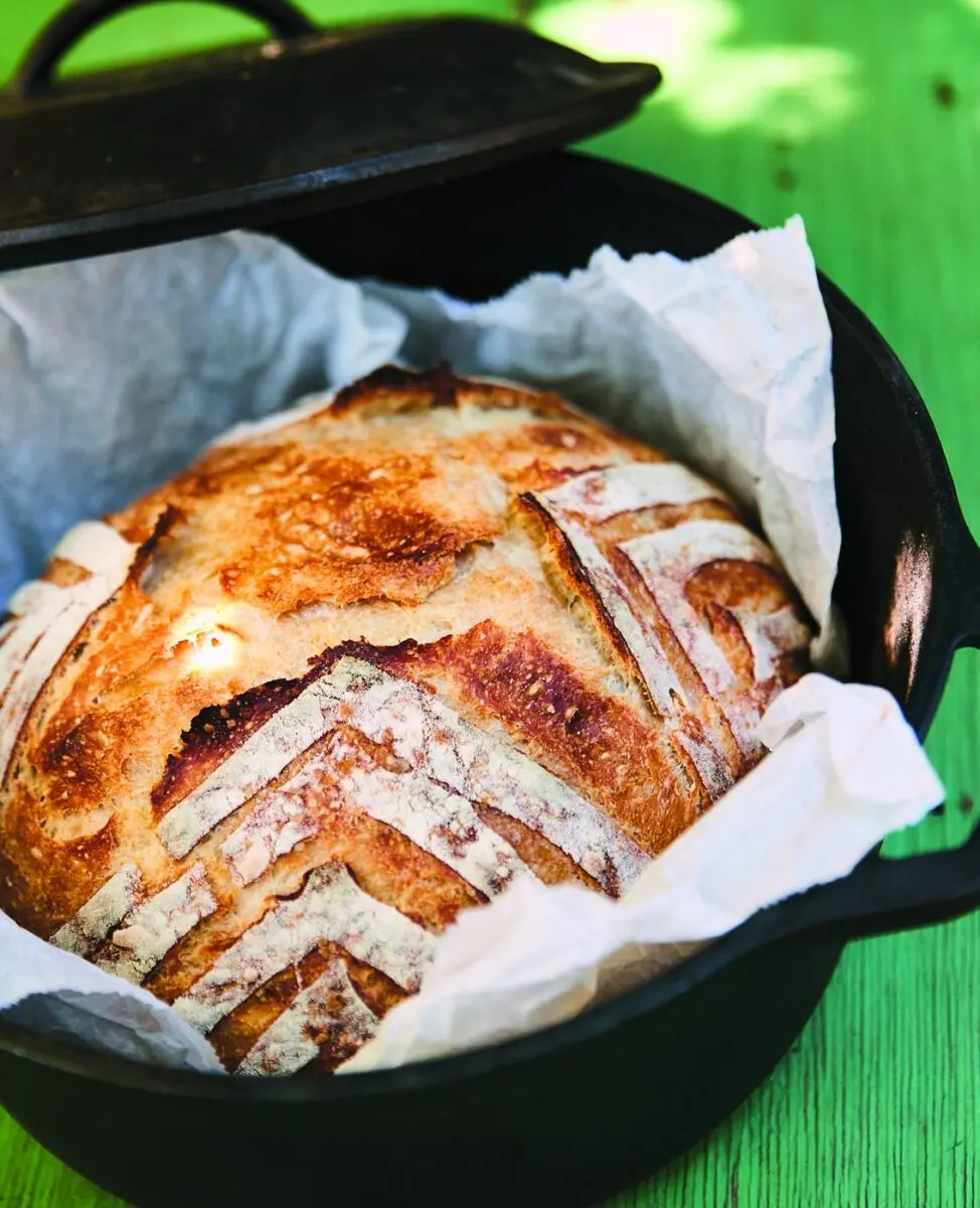 Dutch Oven Bread 1 Golden dutch oven bread with a scored crust lifting out of a cast iron pot on parchment